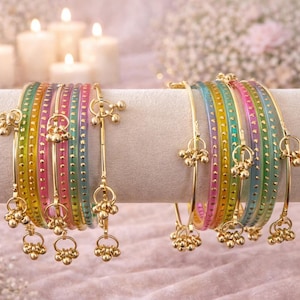 Può includere: Braccialetti colorati con dettagli dorati e ciondoli pendenti. I braccialetti sono disposti in set, mostrando uno spettro di colori tra cui rosa, verde e giallo. Lo sfondo presenta luci soffuse e candele.