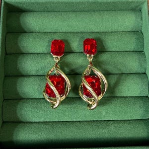 May include: A pair of gold-tone drop earrings featuring a rectangular red faceted stone at the top and a larger matching rectangular red stone suspended within a swirling gold-tone metal cage design, creating a layered and dimensional appearance.