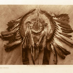 Peut inclure: Photographie sépia d'un bouclier Arapaho. Le bouclier est circulaire, avec un motif central et orné de plumes et de glands suspendus. L'image est une photographie historique, avec le texte "ARAPAHO SHIELD" en bas.