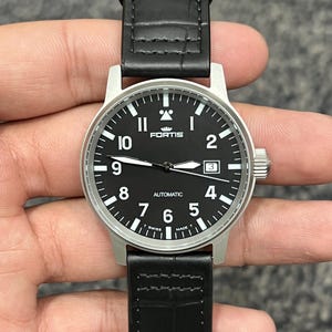 May include: A black watch with a black leather strap. The watch face is black with white numerals and hands. The watch has the word "FORTIS" at the top and "AUTOMATIC" at the bottom. The watch is Swiss made.