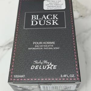 May include: Black Dusk Eau de Toilette spray for men. The box is black with a textured pattern and pink stitching. The text on the box includes "POUR HOMME", "EAU DE TOILETTE", and "Shirley May Deluxe". The bottle contains 3.4 fl oz (100ml).