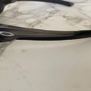 May include: Dark gray sunglasses with a curved frame and dark lenses. The sunglasses have a silver emblem on the side and are set against a light-colored, marble-like surface.
