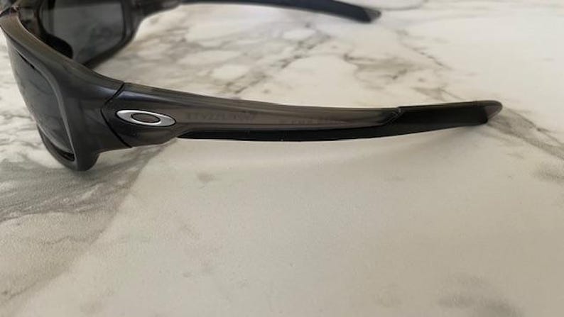 May include: Dark gray sunglasses with a curved frame and dark lenses. The sunglasses have a silver emblem on the side and are set against a light-colored, marble-like surface.