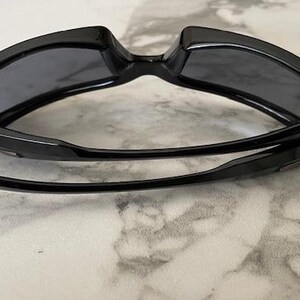 K&ouml;nnte beinhalten: Schwarze Sonnenbrille mit dunkelgrauen Gl&auml;sern. Die Sonnenbrille hat ein schlankes, modernes Design mit einem dicken Rahmen und gebogenen B&uuml;geln. Die Gl&auml;ser sind rechteckig und bieten volle Abdeckung. Die Sonnenbrille ist zusammengeklappt.