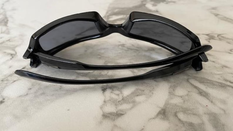 K&ouml;nnte beinhalten: Schwarze Sonnenbrille mit dunkelgrauen Gl&auml;sern. Die Sonnenbrille hat ein schlankes, modernes Design mit einem dicken Rahmen und gebogenen B&uuml;geln. Die Gl&auml;ser sind rechteckig und bieten volle Abdeckung. Die Sonnenbrille ist zusammengeklappt.