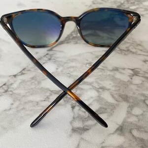May include: Sunglasses with a tortoiseshell pattern frame and blue-tinted lenses. The arms of the sunglasses are crossed, showcasing the design. The lenses reflect a gradient of blue and green hues.