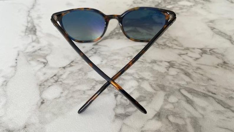 May include: Sunglasses with a tortoiseshell pattern frame and blue-tinted lenses. The arms of the sunglasses are crossed, showcasing the design. The lenses reflect a gradient of blue and green hues.
