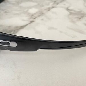Puede incluir: Gafas de sol negras con un dise&ntilde;o elegante y moderno. La montura es negra con un detalle plateado en la patilla. Las lentes tienen un degradado de amarillo a azul. Las gafas de sol est&aacute;n sobre una superficie de m&aacute;rmol blanco y gris.