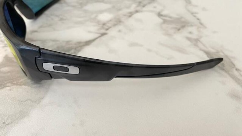 Puede incluir: Gafas de sol negras con un dise&ntilde;o elegante y moderno. La montura es negra con un detalle plateado en la patilla. Las lentes tienen un degradado de amarillo a azul. Las gafas de sol est&aacute;n sobre una superficie de m&aacute;rmol blanco y gris.