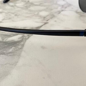 May include: Black sunglasses with a dark blue frame and gray lenses. The sunglasses have a sleek, modern design with a curved temple arm. The Oakley logo is visible on the temple.