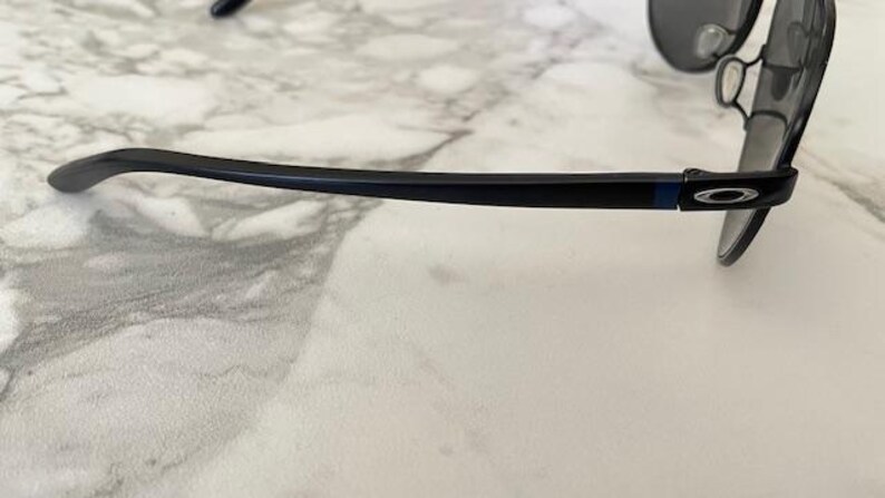 May include: Black sunglasses with a dark blue frame and gray lenses. The sunglasses have a sleek, modern design with a curved temple arm. The Oakley logo is visible on the temple.