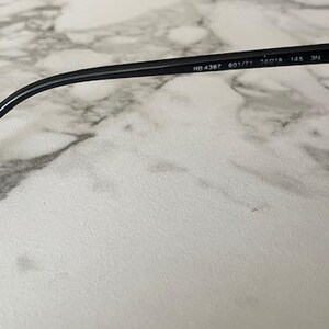 Puede incluir: Gafas de sol negras con lentes oscuras y montura negra. La patilla de las gafas es visible, con el texto "RB 4387 601/71 56[]18 145 3N" impreso en la patilla. Las gafas est&aacute;n sobre una superficie de m&aacute;rmol.