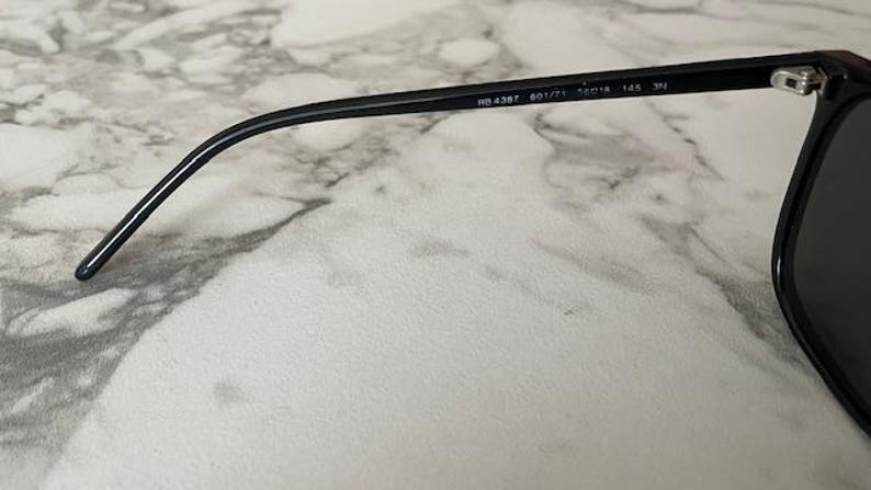 Puede incluir: Gafas de sol negras con lentes oscuras y montura negra. La patilla de las gafas es visible, con el texto "RB 4387 601/71 56[]18 145 3N" impreso en la patilla. Las gafas est&aacute;n sobre una superficie de m&aacute;rmol.