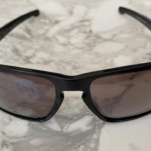 May include: Black sunglasses with dark lenses. The sunglasses have a rectangular frame and a sleek, modern design. The arms of the sunglasses are thin and extend back to the ears. The lenses are a dark, smoky color.