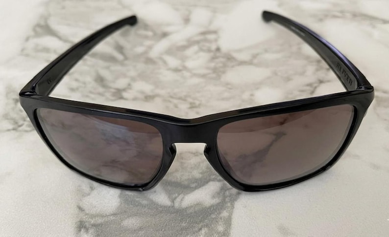 May include: Black sunglasses with dark lenses. The sunglasses have a rectangular frame and a sleek, modern design. The arms of the sunglasses are thin and extend back to the ears. The lenses are a dark, smoky color.