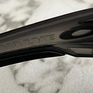 May include: Close-up of a black sunglass arm with the word "VALYTE" embossed on the side. The sunglasses appear to be made of a durable, matte material. The image is taken against a blurred, light-coloured background.