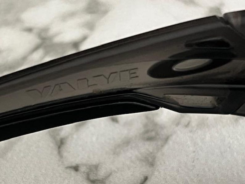 May include: Close-up of a black sunglass arm with the word "VALYTE" embossed on the side. The sunglasses appear to be made of a durable, matte material. The image is taken against a blurred, light-coloured background.