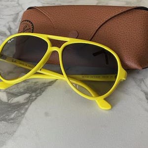 May include: Bright yellow sunglasses with dark lenses and a matching case. The sunglasses have a classic aviator shape with a double bridge and a curved brow bar. The case is brown and made of a textured material.