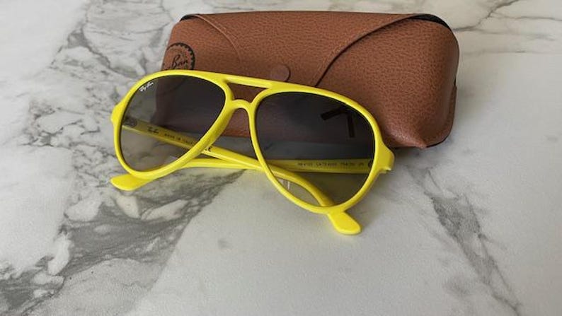 May include: Bright yellow sunglasses with dark lenses and a matching case. The sunglasses have a classic aviator shape with a double bridge and a curved brow bar. The case is brown and made of a textured material.