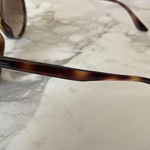 May include: Brown tortoiseshell sunglasses with dark lenses. The sunglasses have a classic design with a thick frame and arms. The Ray-Ban logo is visible on the arm. The sunglasses are resting on a white and gray marble surface.