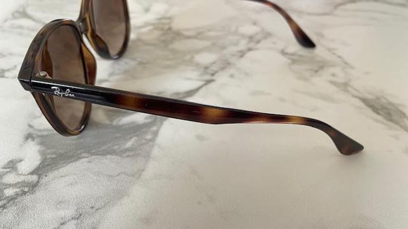 May include: Brown tortoiseshell sunglasses with dark lenses. The sunglasses have a classic design with a thick frame and arms. The Ray-Ban logo is visible on the arm. The sunglasses are resting on a white and gray marble surface.
