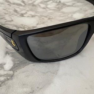 May include: Black sunglasses with a rectangular frame and dark lenses. The sunglasses have a curved design and a small gold emblem on the side. The lenses reflect the light, and the frame appears to be made of plastic.