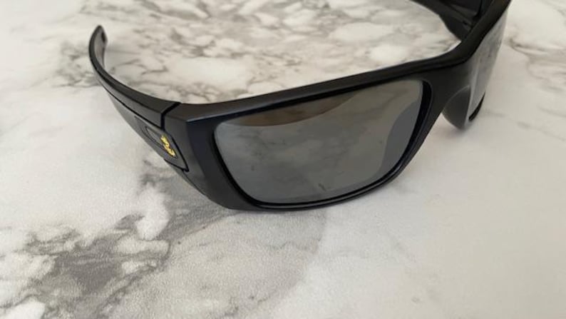 May include: Black sunglasses with a rectangular frame and dark lenses. The sunglasses have a curved design and a small gold emblem on the side. The lenses reflect the light, and the frame appears to be made of plastic.