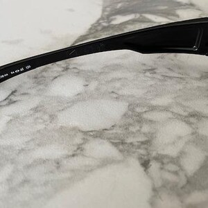 K&ouml;nnte beinhalten: Nahaufnahme einer schwarzen Sonnenbrille mit rechteckigem Rahmen und dunklen Gl&auml;sern. Der B&uuml;gel tr&auml;gt den Text "OO9238-04 54 18 130". Die Sonnenbrille liegt auf einer hellen, marmorierten Oberfl&auml;che.