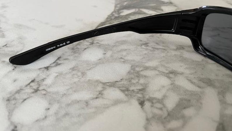 K&ouml;nnte beinhalten: Nahaufnahme einer schwarzen Sonnenbrille mit rechteckigem Rahmen und dunklen Gl&auml;sern. Der B&uuml;gel tr&auml;gt den Text "OO9238-04 54 18 130". Die Sonnenbrille liegt auf einer hellen, marmorierten Oberfl&auml;che.