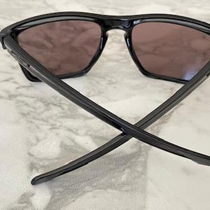 May include: Black sunglasses with a square frame and dark lenses. The arms of the sunglasses are crossed, resting on a marble-patterned surface. The sunglasses are a sleek, modern design.
