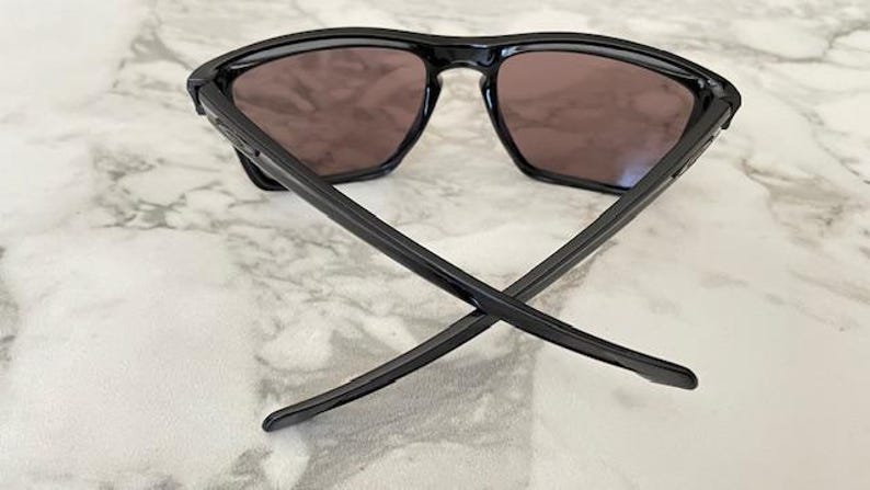 May include: Black sunglasses with a square frame and dark lenses. The arms of the sunglasses are crossed, resting on a marble-patterned surface. The sunglasses are a sleek, modern design.