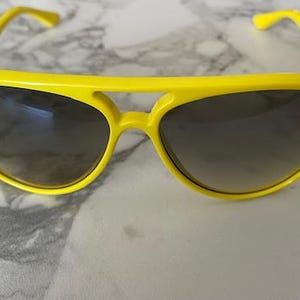 May include: Bright yellow sunglasses with dark lenses. The sunglasses have a classic aviator style with a thick, solid frame. The Ray-Ban logo is visible on the left lens. The sunglasses are resting on a marble surface.