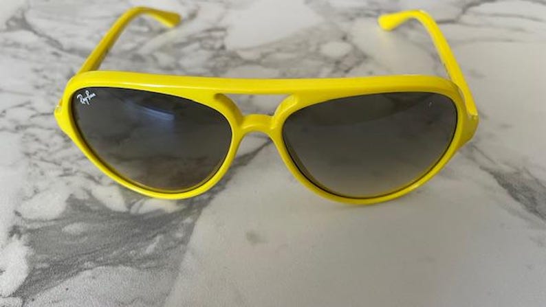 May include: Bright yellow sunglasses with dark lenses. The sunglasses have a classic aviator style with a thick, solid frame. The Ray-Ban logo is visible on the left lens. The sunglasses are resting on a marble surface.
