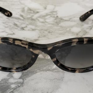 Puede incluir: Un par de gafas de sol Ray-Ban con un patr&oacute;n de carey. Las gafas de sol tienen una forma de ojo de gato cl&aacute;sica con lentes oscuras y el logotipo de Ray-Ban en blanco en la lente izquierda.