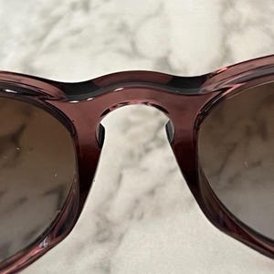 May include: Close-up of a pair of sunglasses with a dark red frame and brown lenses. The sunglasses have a classic, rounded shape. The image is well-lit, highlighting the details of the frame and lenses.