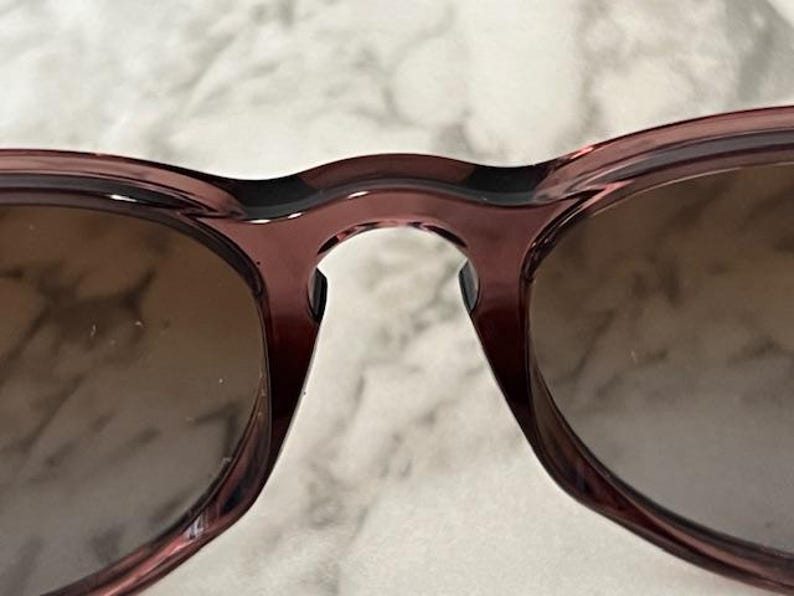 May include: Close-up of a pair of sunglasses with a dark red frame and brown lenses. The sunglasses have a classic, rounded shape. The image is well-lit, highlighting the details of the frame and lenses.