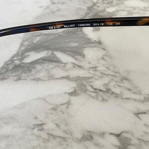 May include: Close-up of sunglasses with a tortoiseshell pattern on the frame. The sunglasses have blue lenses and the text "RB 2157 ELLIOT 1356/3M 52o19 145 2N" is printed on the arm.