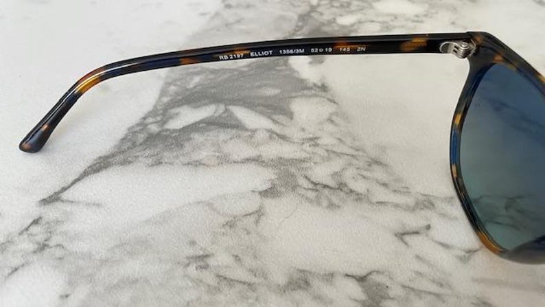 May include: Close-up of sunglasses with a tortoiseshell pattern on the frame. The sunglasses have blue lenses and the text "RB 2157 ELLIOT 1356/3M 52o19 145 2N" is printed on the arm.