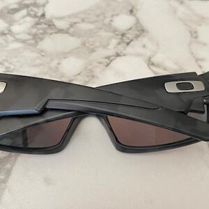 Puede incluir: Gafas de sol negras con un dise&ntilde;o geom&eacute;trico. Las gafas tienen montura y lentes oscuras. El logotipo de Oakley es visible en las patillas. Las gafas est&aacute;n plegadas.