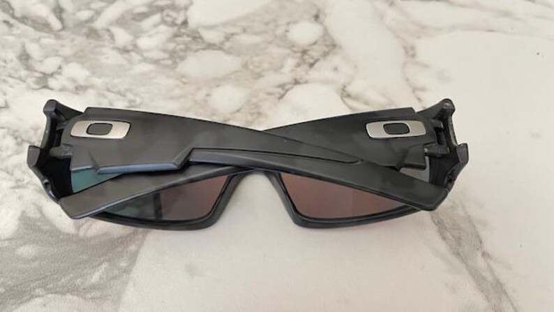 Puede incluir: Gafas de sol negras con un dise&ntilde;o geom&eacute;trico. Las gafas tienen montura y lentes oscuras. El logotipo de Oakley es visible en las patillas. Las gafas est&aacute;n plegadas.