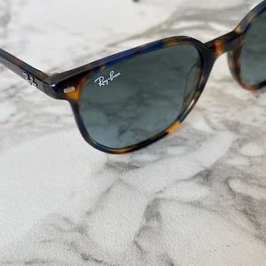 May include: Tortoiseshell sunglasses with dark lenses. The frames have a brown and yellow pattern with blue accents. The Ray-Ban logo is visible on the lens. The sunglasses are on a white marble surface.