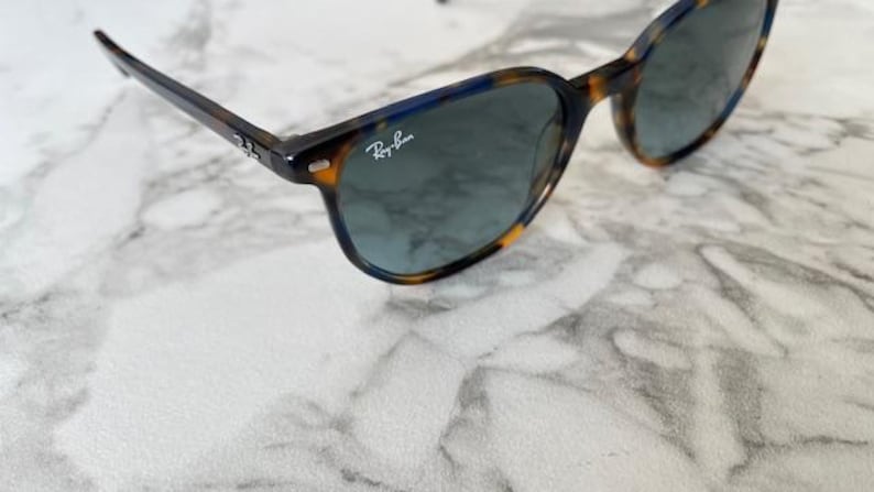 May include: Tortoiseshell sunglasses with dark lenses. The frames have a brown and yellow pattern with blue accents. The Ray-Ban logo is visible on the lens. The sunglasses are on a white marble surface.