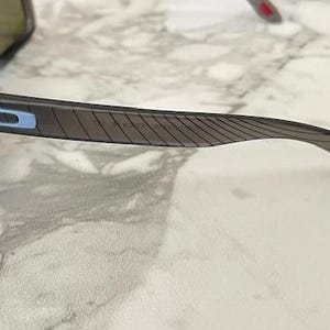 May include: Dark gray sunglasses with a black frame. The temples have a textured design and a blue and white logo. The lenses are a dark green color.