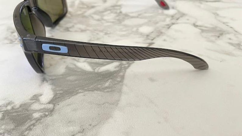 May include: Dark gray sunglasses with a black frame. The temples have a textured design and a blue and white logo. The lenses are a dark green color.