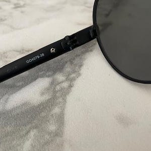 May include: Black aviator sunglasses with dark lenses. The frame is black and the arm of the glasses has the number "004079-38" printed on it. The sunglasses are on a white and gray surface.