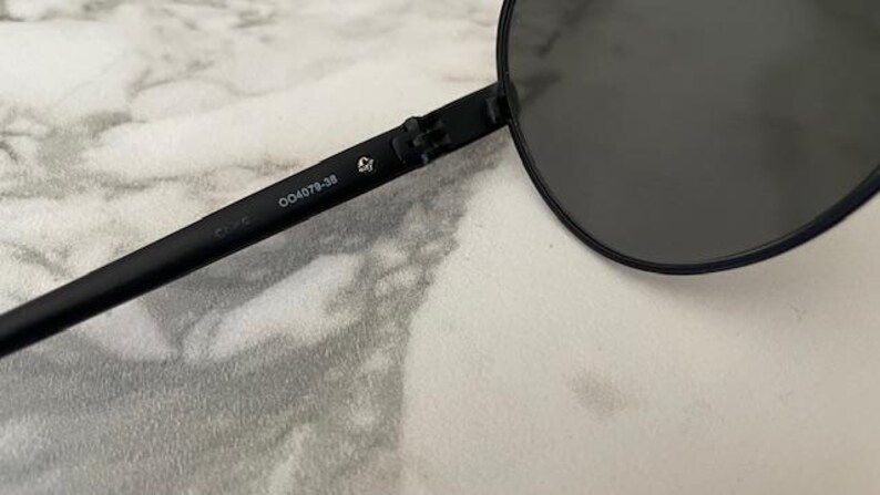 May include: Black aviator sunglasses with dark lenses. The frame is black and the arm of the glasses has the number "004079-38" printed on it. The sunglasses are on a white and gray surface.
