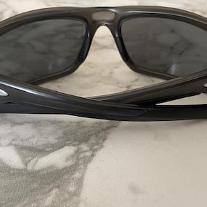 May include: Black sunglasses with dark tinted lenses. The sunglasses have a curved frame and arms, with the Oakley logo visible on the temples. The sunglasses are folded, resting on a light-colored surface.