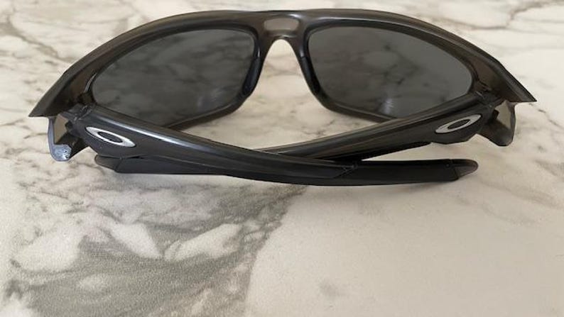 May include: Black sunglasses with dark tinted lenses. The sunglasses have a curved frame and arms, with the Oakley logo visible on the temples. The sunglasses are folded, resting on a light-colored surface.