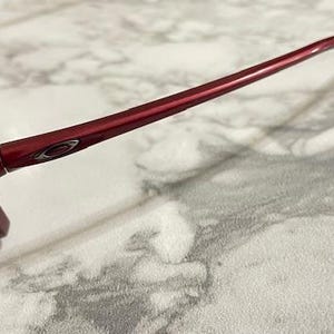 May include: Close-up of a pair of red sunglasses. The sunglasses have a glossy finish and a sleek design. The frame is a deep red color, and the lenses are a dark tint. The Oakley logo is visible on the temple.