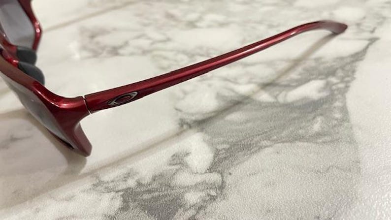 May include: Close-up of a pair of red sunglasses. The sunglasses have a glossy finish and a sleek design. The frame is a deep red color, and the lenses are a dark tint. The Oakley logo is visible on the temple.